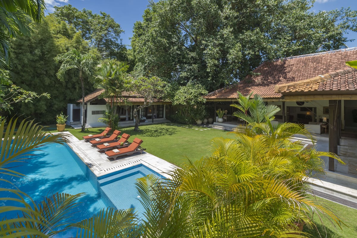 A pool with lounge chairs, surrounded by lush greenery and a building with a tiled roof.