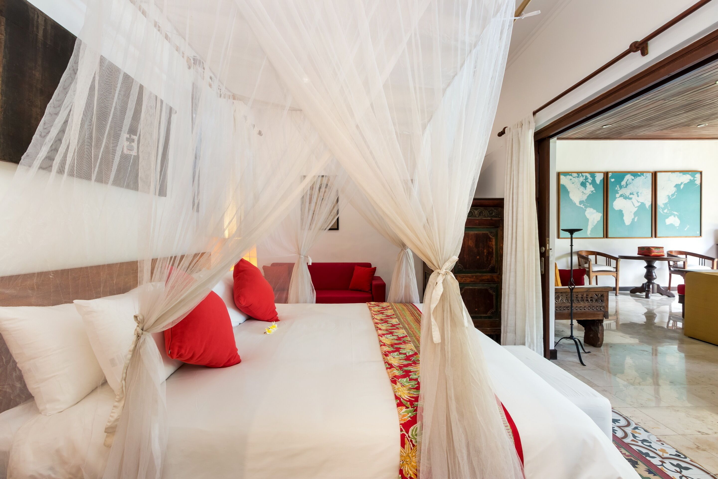 Un lit à baldaquin, des oreillers rouges et une couvre-lit à motifs floraux. On aperçoit un canapé rouge à l’arrière-plan et une carte du monde accrochée au mur.