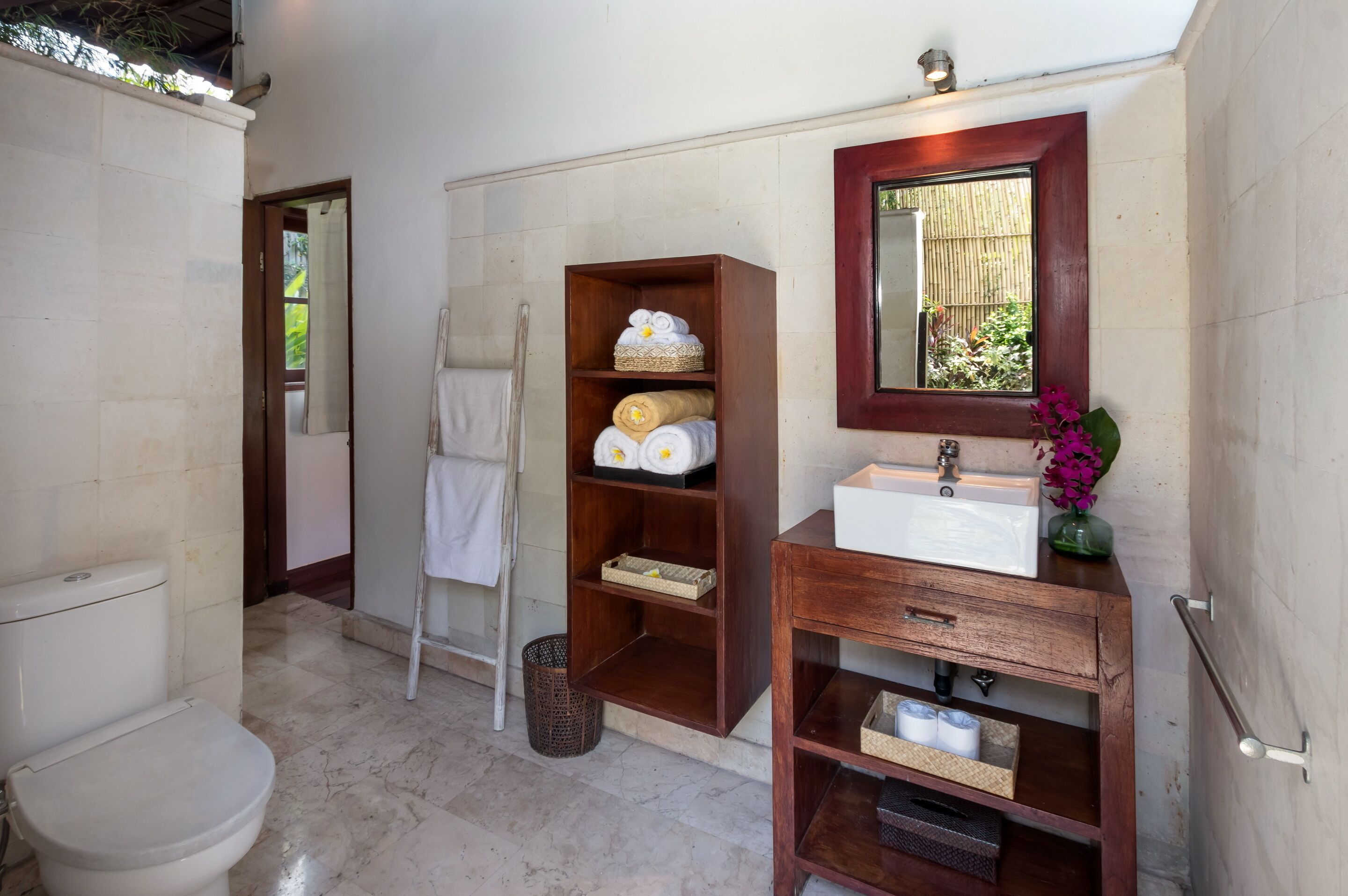 A bathroom with a toilet, a wooden shelf with towels, a sink with a mirror, and a small window with a view of greenery.
