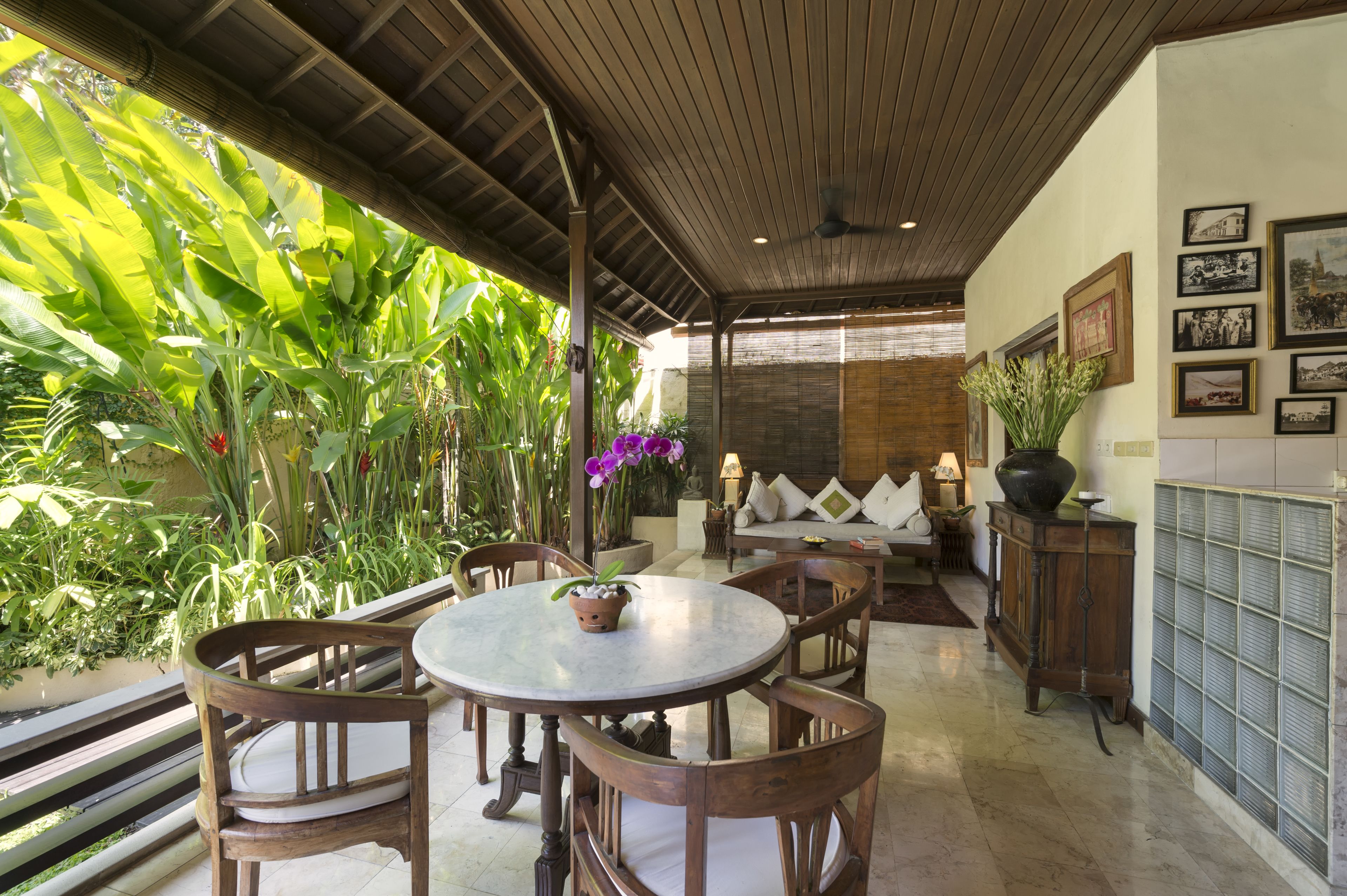 A covered patio with a round table and chairs, surrounded by lush tropical plants and a sofa with cushions.