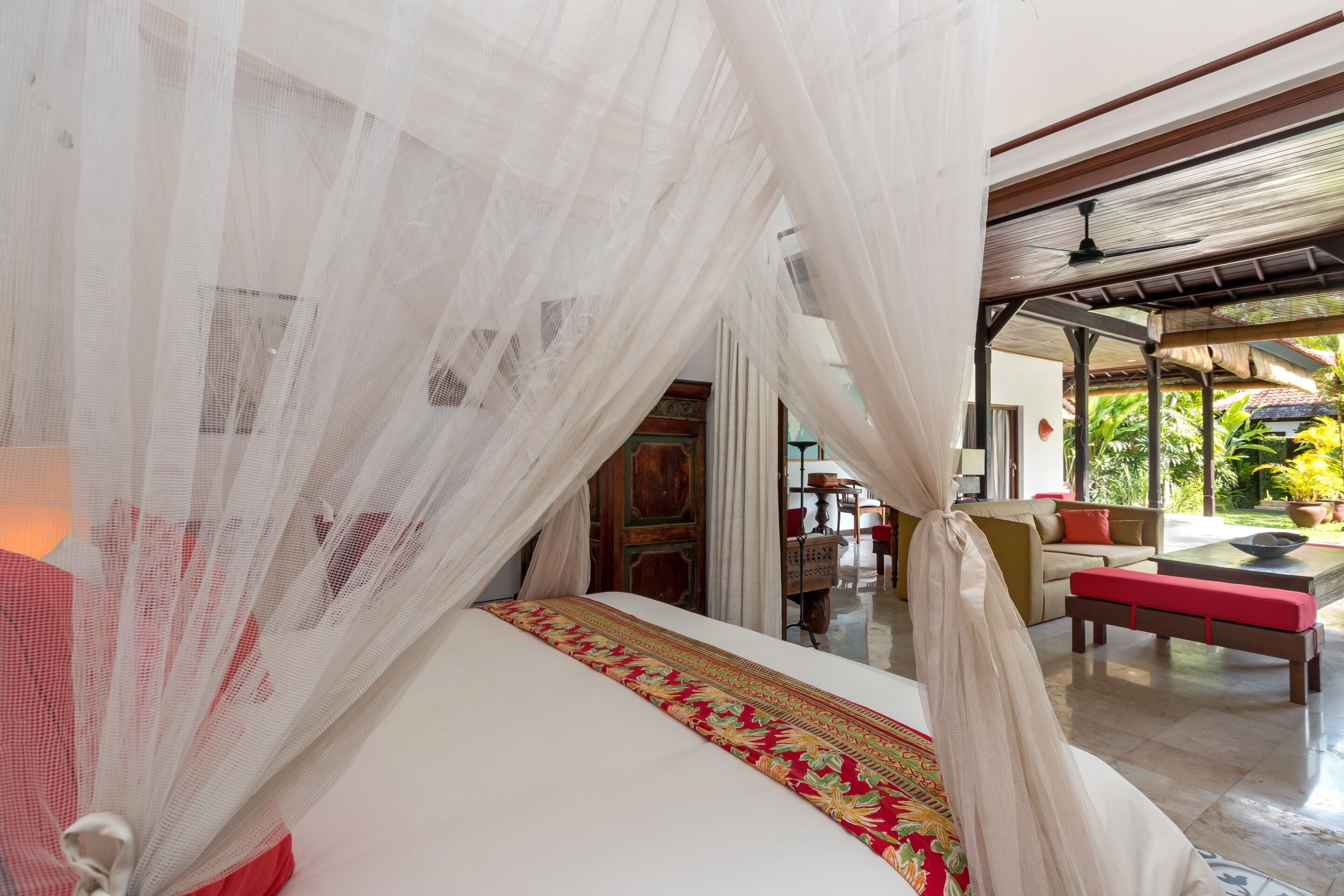 A room with a bed covered by a canopy, a red and gold patterned bedspread, and a view of an outdoor seating area with a table and chairs.