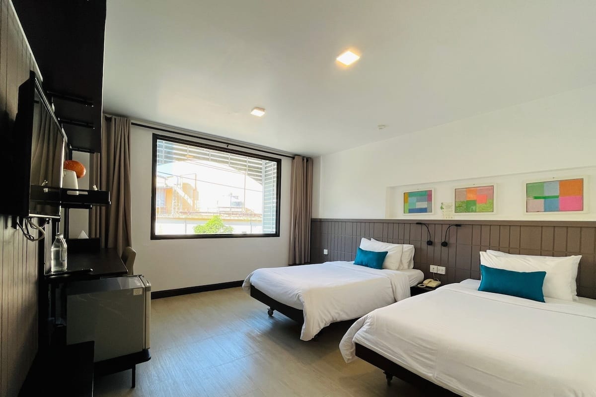 Quarto de hotel com duas camas, uma escrivaninha, uma cadeira e uma janela com persianas.