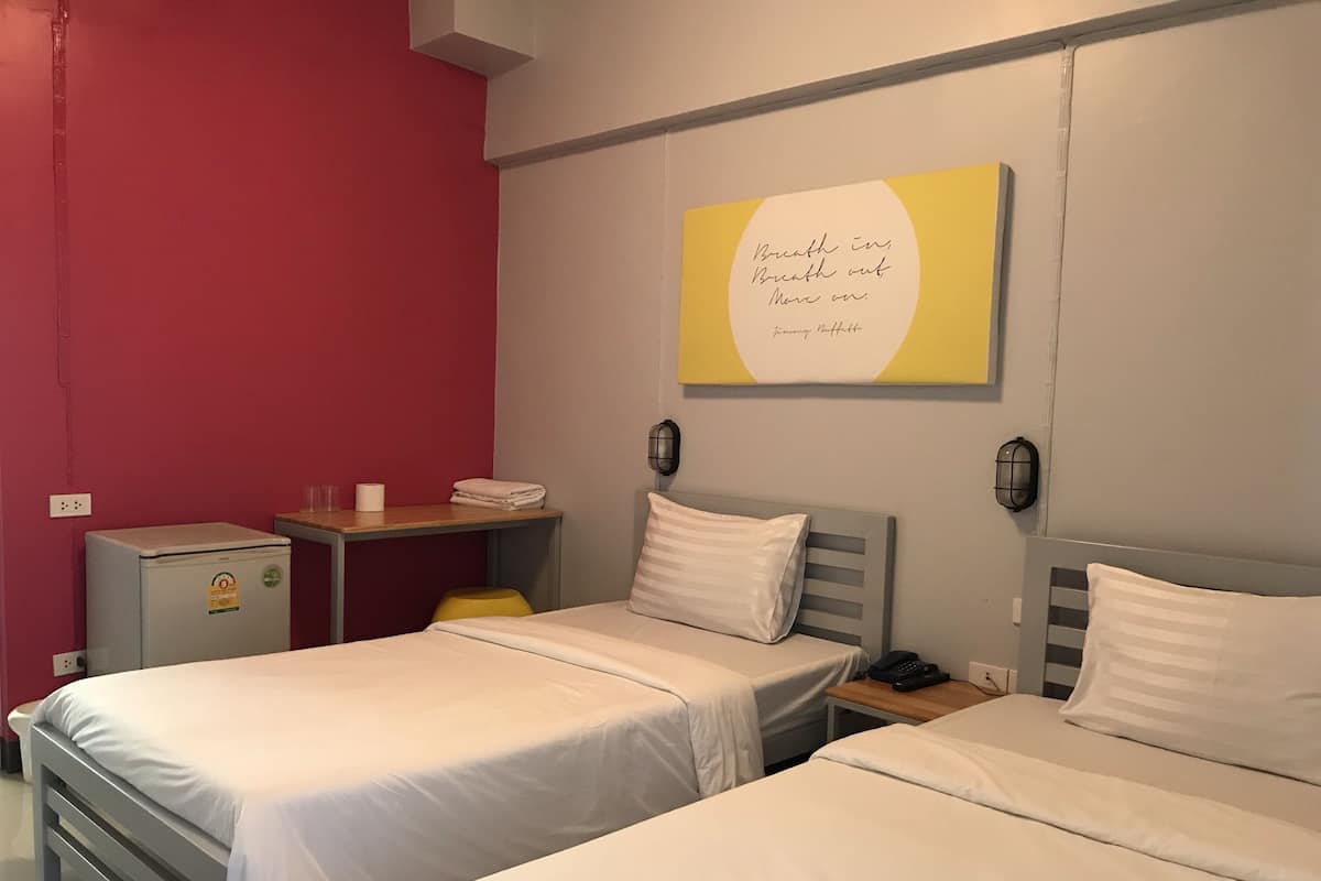 A hotel room with two single beds, a small desk with a phone, a refrigerator, and a wall-mounted light fixture.