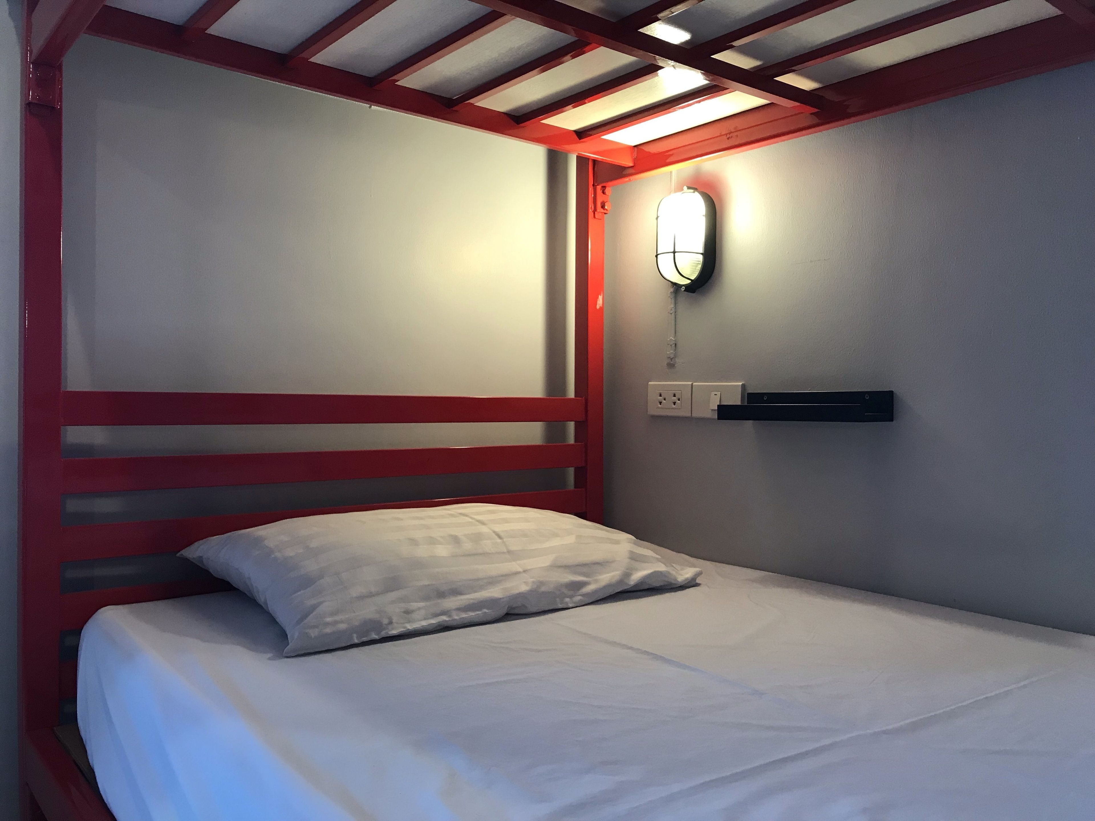 A single bed with a red headboard, white bedding, a wall-mounted light, and a power outlet.