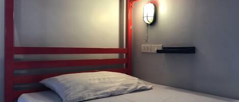 A single bed with a red headboard, white bedding, a wall-mounted light, and a power outlet.