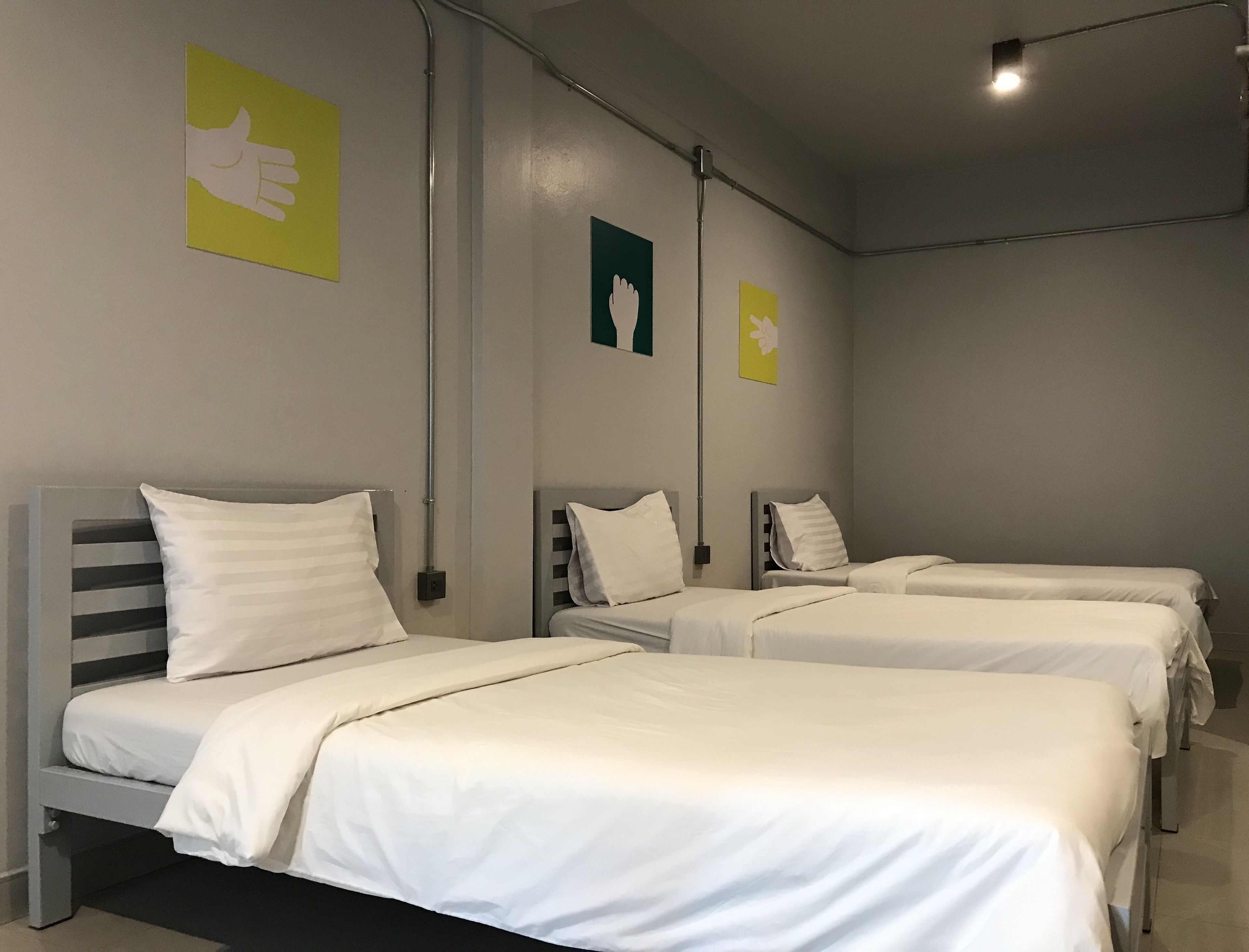 A hotel room with four single beds, each with white bedding and a grey headboard. There are two wall-mounted light fixtures and a ceiling light.