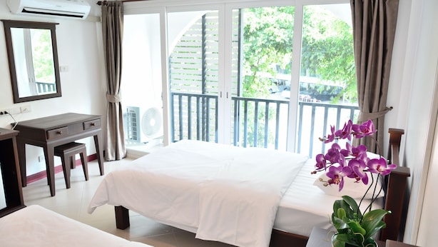 A hotel room with a bed, a desk, a chair, a potted plant, and a balcony with a view of greenery.
