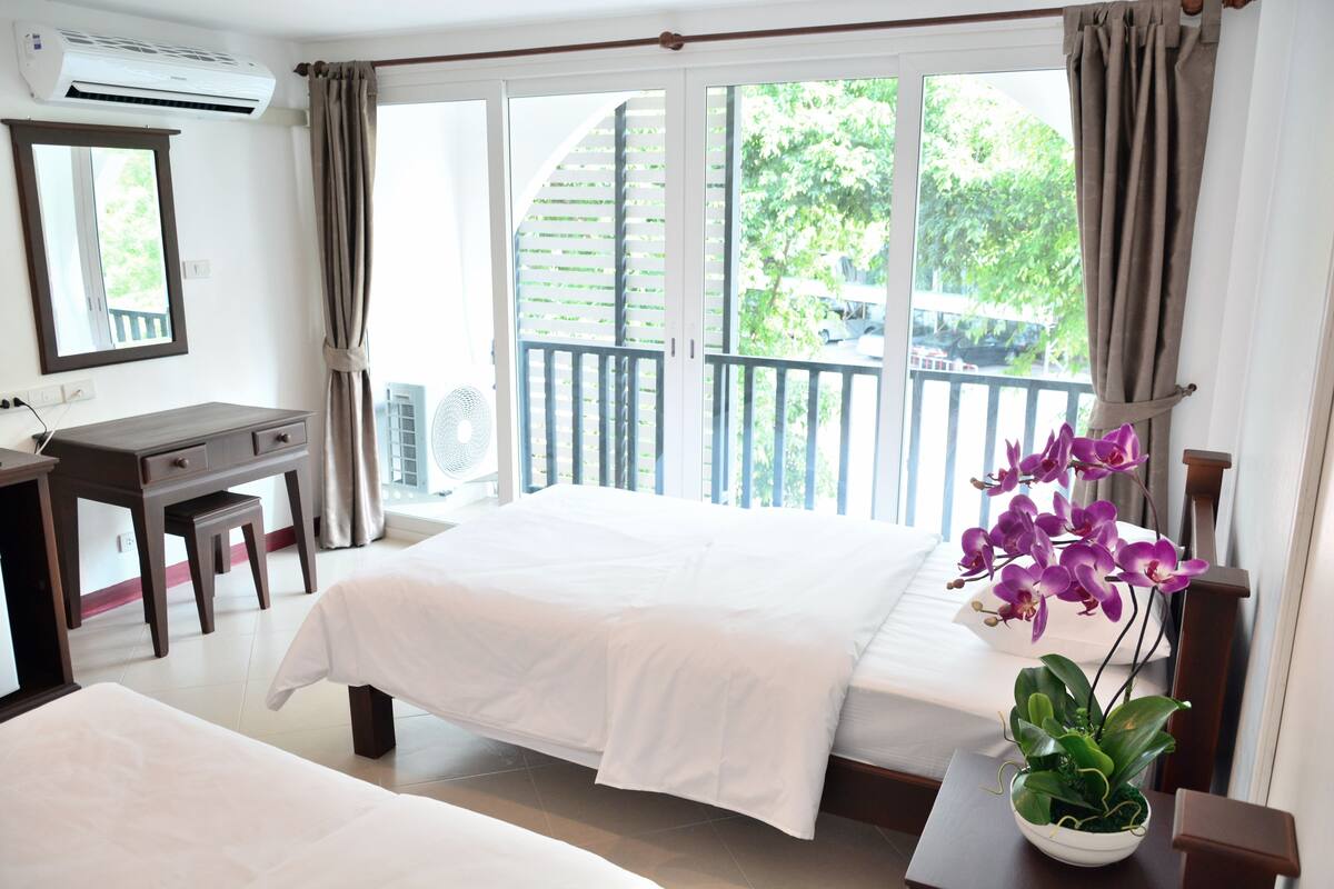 Quarto de hotel com cama, escrivaninha, cadeira, planta em vaso e varanda com vista para áreas verdes.
