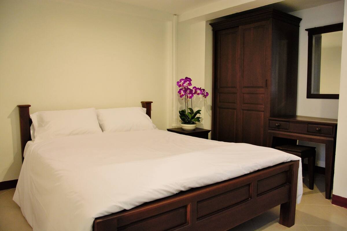 Quarto com uma cama grande, uma mesa de cabeceira de madeira, uma orquídea em vaso, um guarda-roupa de madeira escura e uma pequena escrivaninha com gavetas.