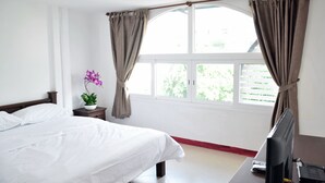 A hotel room with a bed, a desk with a computer, a window with curtains, and a potted plant.