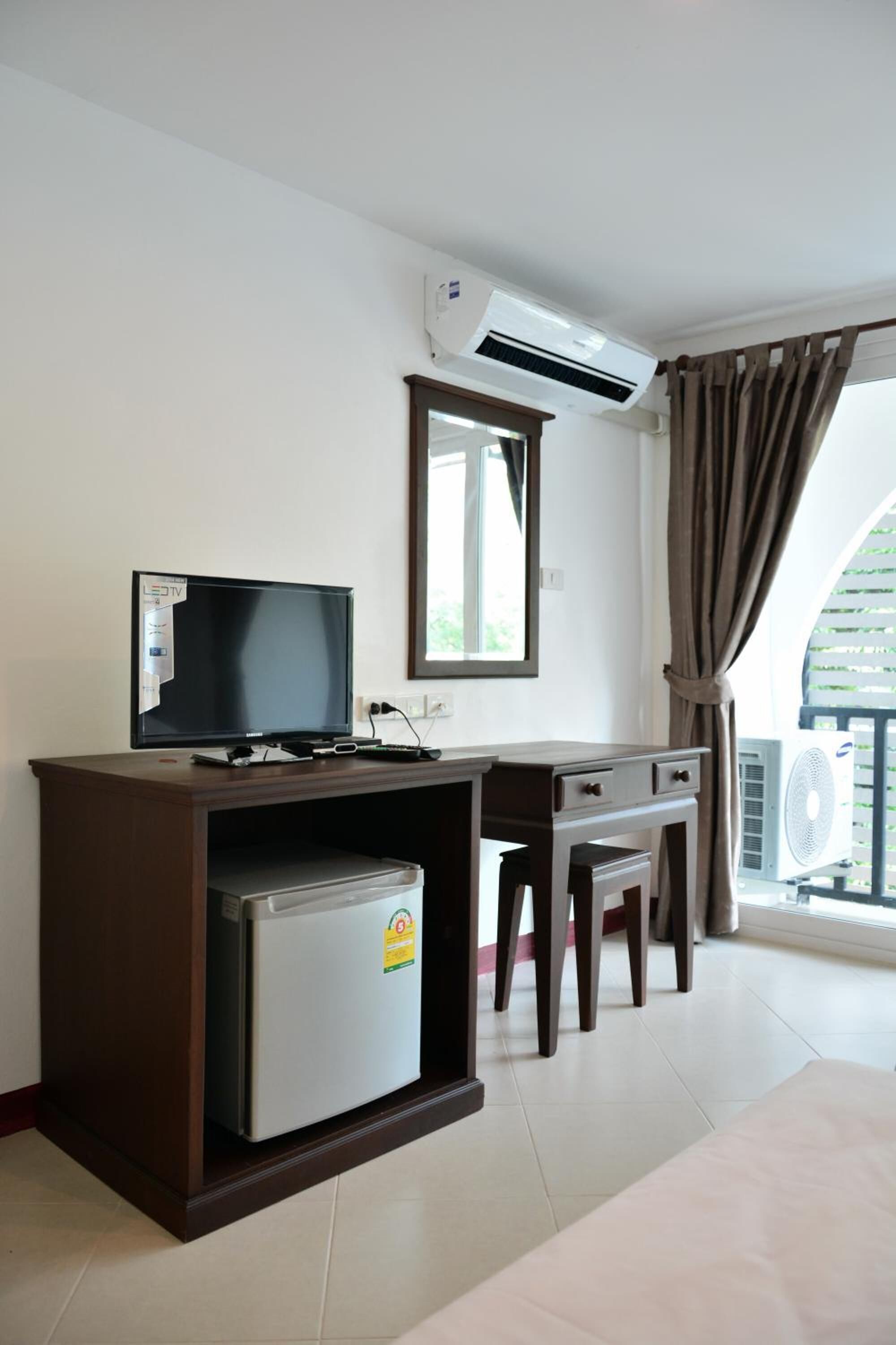 A room with a TV, a small refrigerator, a desk, a chair, an air conditioning unit, and a window with curtains.