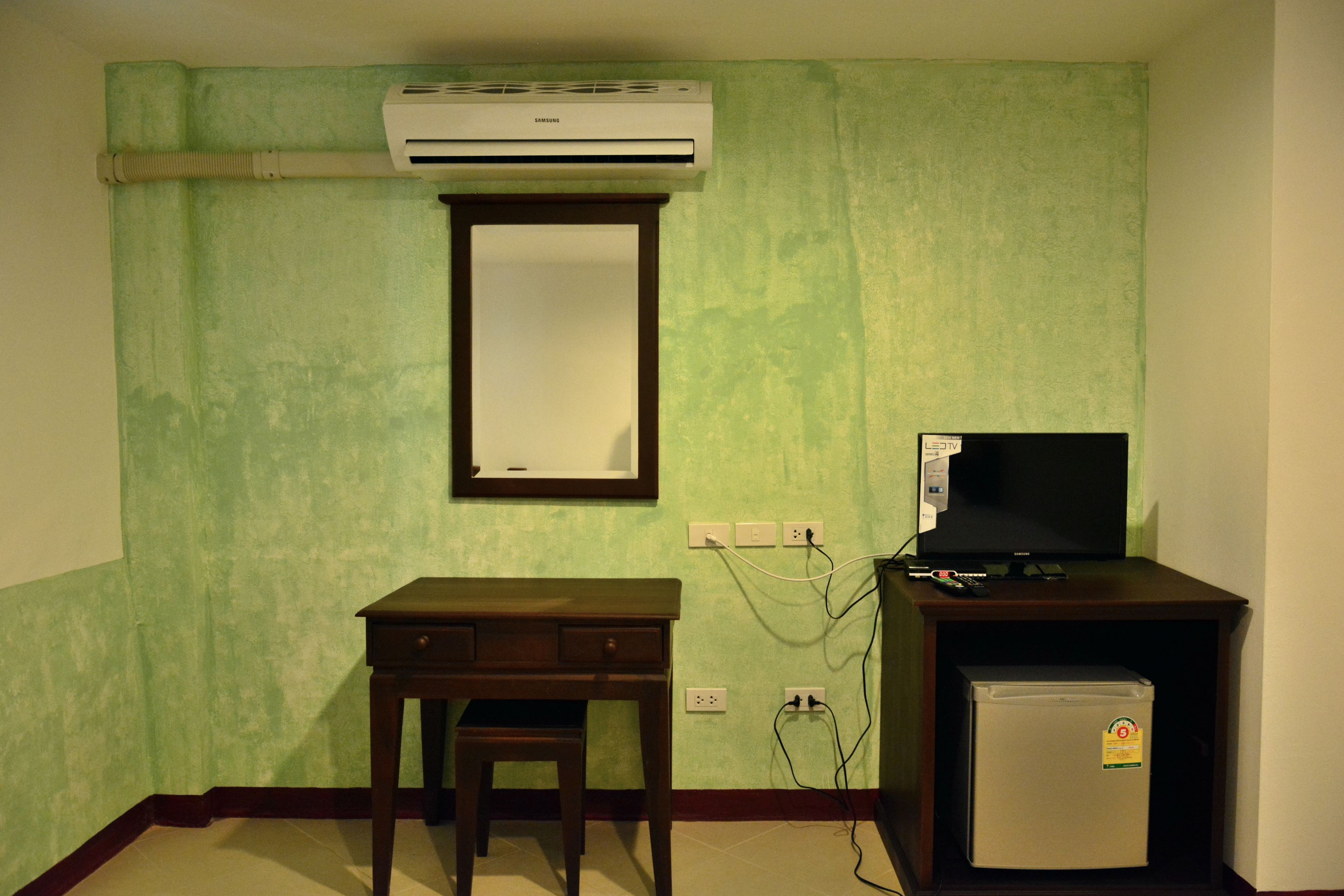 Een kamer met een groene wand, een houten bureau met stoel, een kleine koelkast en een airconditioner boven het bureau.