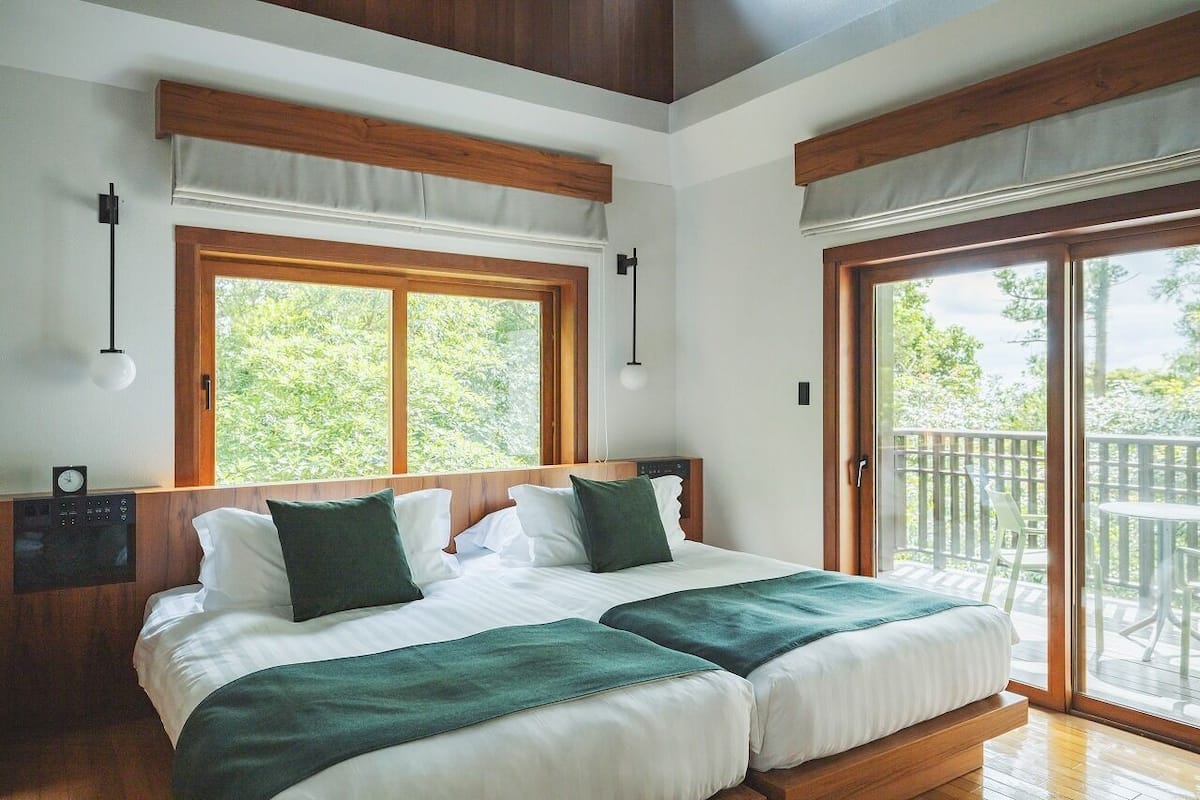 A bedroom with two beds, wooden headboards, large windows, and a balcony with outdoor furniture.