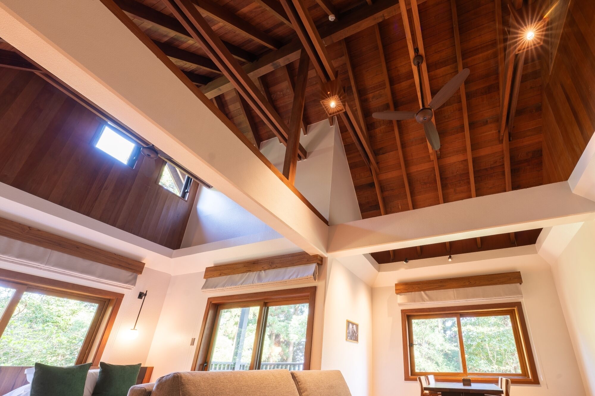 Une pièce dotée d’un plafond haut, de poutres en bois, d’un ventilateur de plafond et de grandes fenêtres offrant une vue sur la verdure.