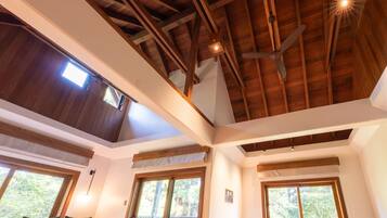 Une pièce dotée d’un plafond haut, de poutres en bois, d’un ventilateur de plafond et de grandes fenêtres offrant une vue sur la verdure.