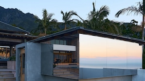 Un bâtiment moderne et minimaliste, doté d’une grande façade en verre, d’une structure en béton et offrant une vue sur l’océan au coucher du soleil.
