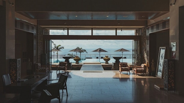Un espace intérieur vaste et lumineux, avec une grande fenêtre donnant sur une piscine et la mer. On y trouve plusieurs chaises et tables disposées pour les repas ou la détente.