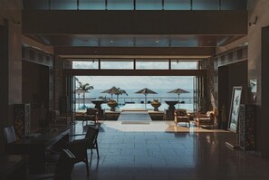 Un espace intérieur vaste et lumineux, avec une grande fenêtre donnant sur une piscine et la mer. On y trouve plusieurs chaises et tables disposées pour les repas ou la détente.