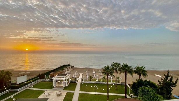 Vista de praia com pôr do sol, cerca branca e palmeiras.