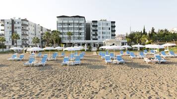 A beach with rows of blue and white sun loungers under white umbrellas, a hotel building in the background, and palm trees.