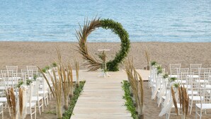 Uma passarela de madeira leva a um espaço para casamento na praia, com cadeiras brancas e plantas.