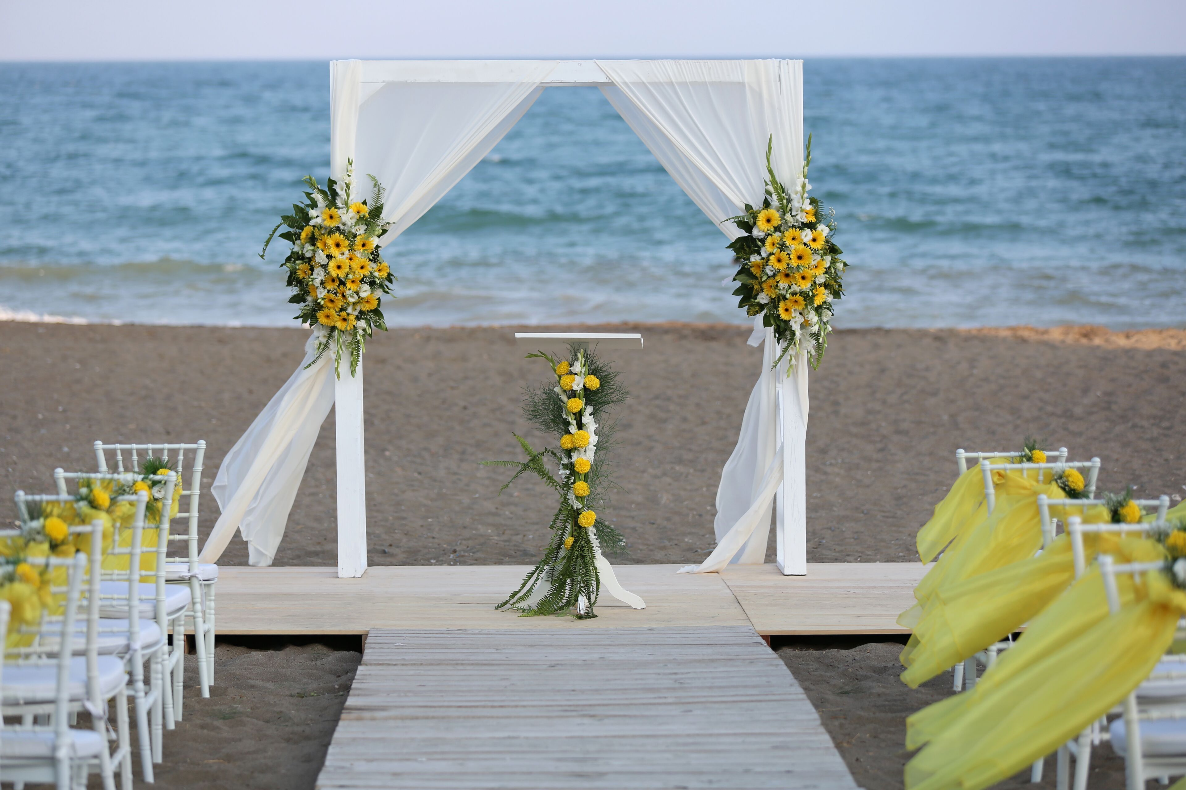 Hochzeitsdekoration am Strand mit einem weißen Bogen, gelben Blumen und weißen Stühlen.