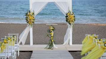 Hochzeitsdekoration am Strand mit einem weißen Bogen, gelben Blumen und weißen Stühlen.