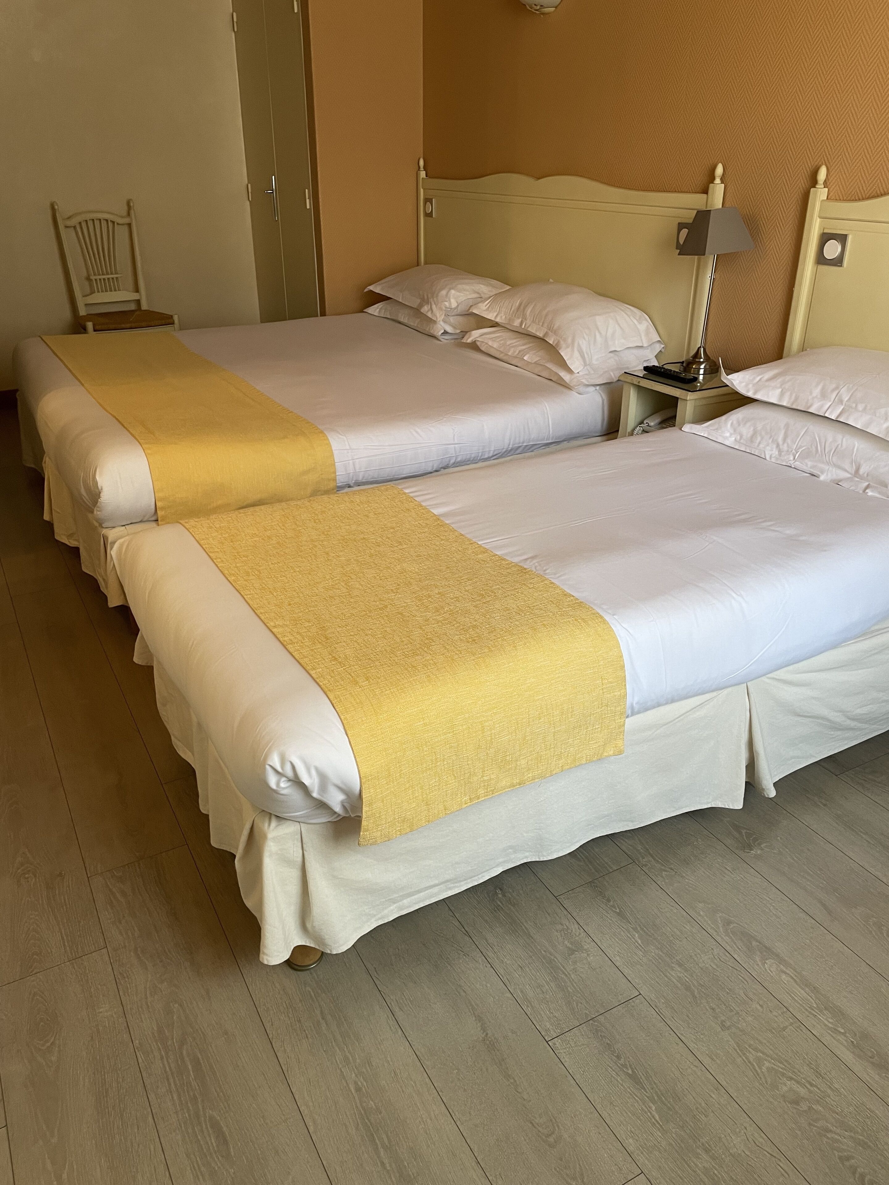 Een hotelkamer met twee bedden, elk voorzien van witte lakens en een gele plaids. Er ligt een houten vloer, een stoel en een nachtkastje met een lamp.