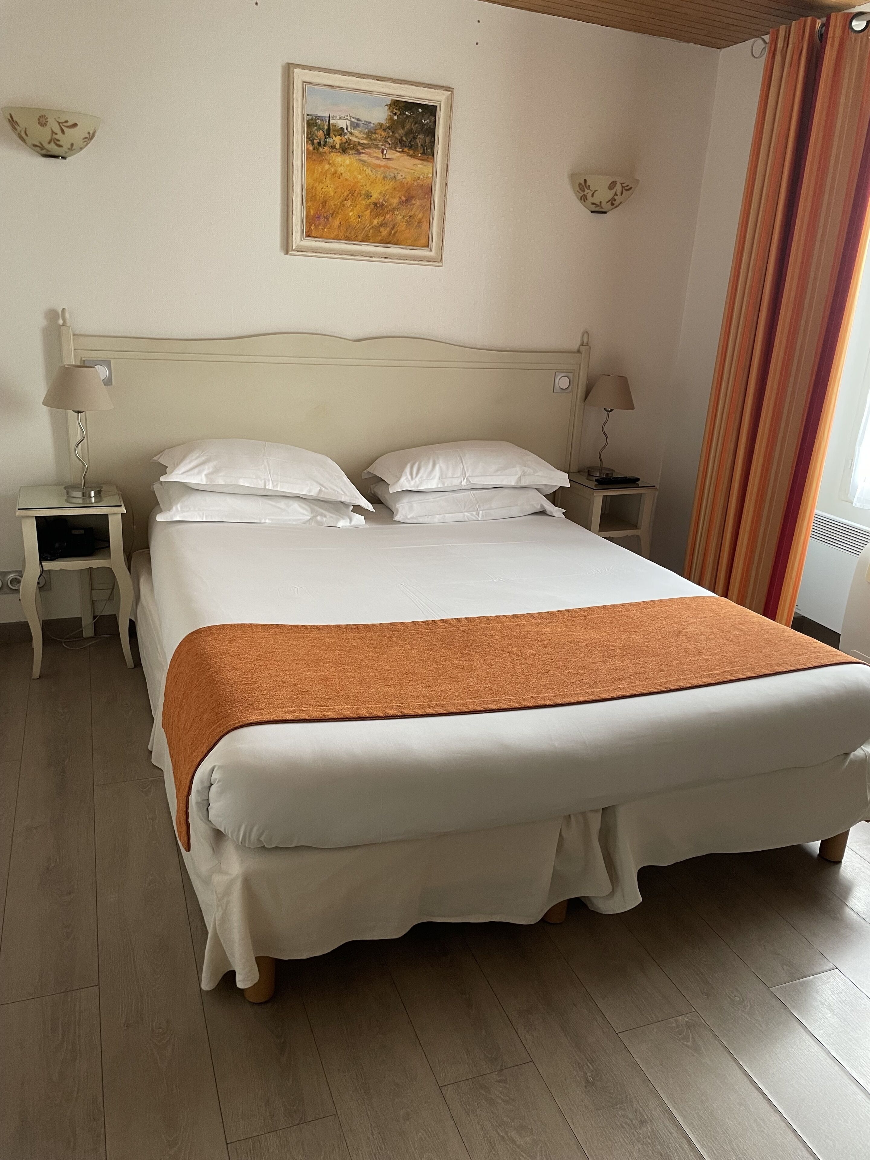A neatly made bed with white linens and a brown throw blanket, a small bedside table with a lamp, and a painting on the wall.