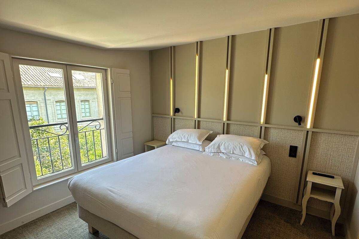 A bedroom with a large bed, a nightstand, a small table, and a view of greenery outside.