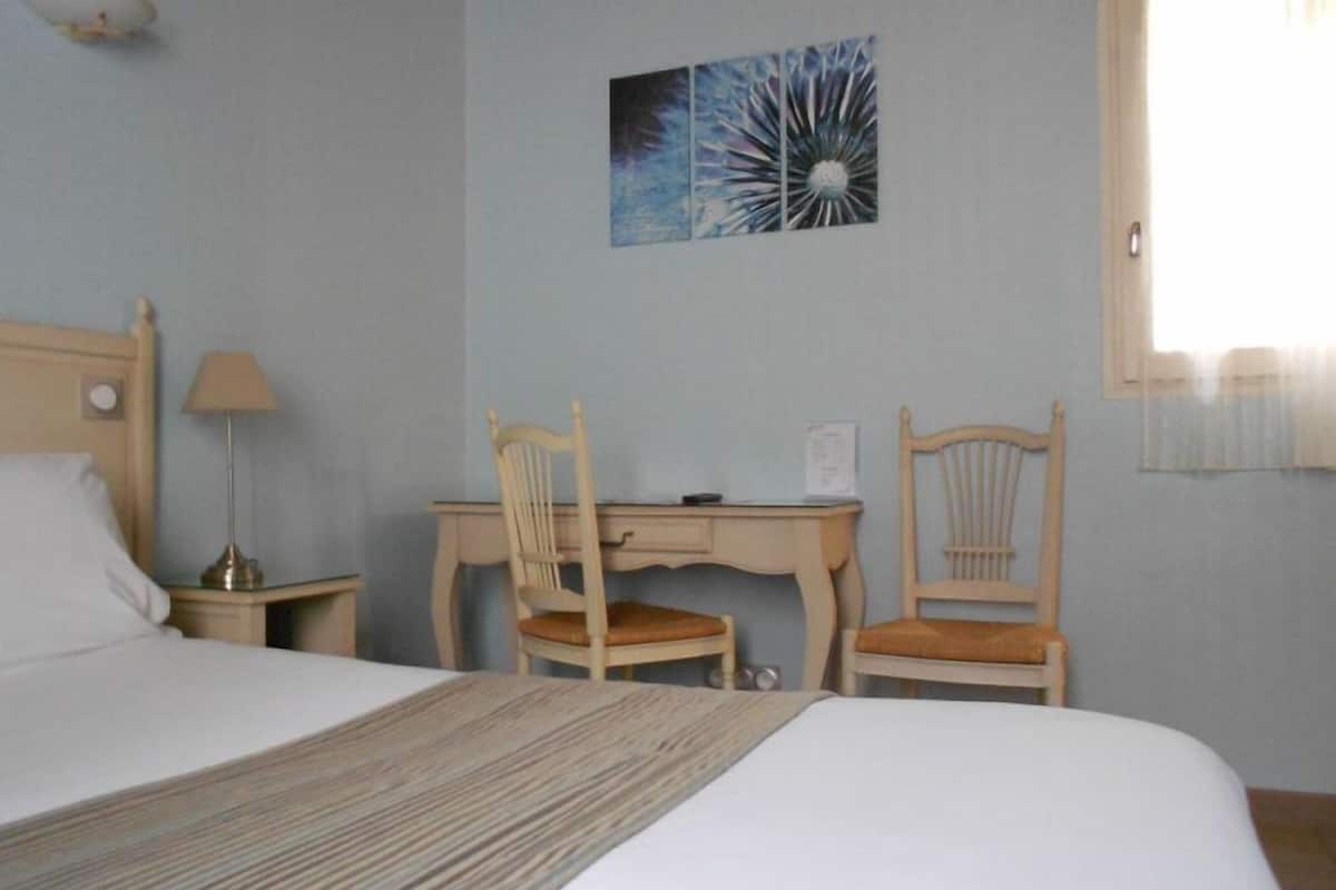 A hotel room with a bed, two wooden chairs, a desk, a lamp, and a painting on the wall.