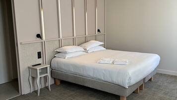 A neatly made bed with white linens and pillows, a small bedside table with a remote control, and a grey carpeted floor.