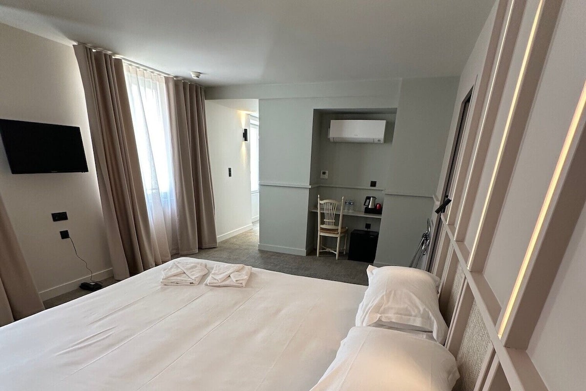 A hotel room with a large bed, a flat-screen TV mounted on the wall, a desk with a chair, and a small kitchenette area.