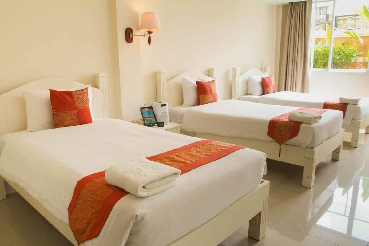 Ein Hotelzimmer mit zwei Betten, jeweils mit weißer Bettwäsche und orangenen Akzenten, einem Nachttisch mit Lampe und einem Fenster mit Blick auf Grünflächen.