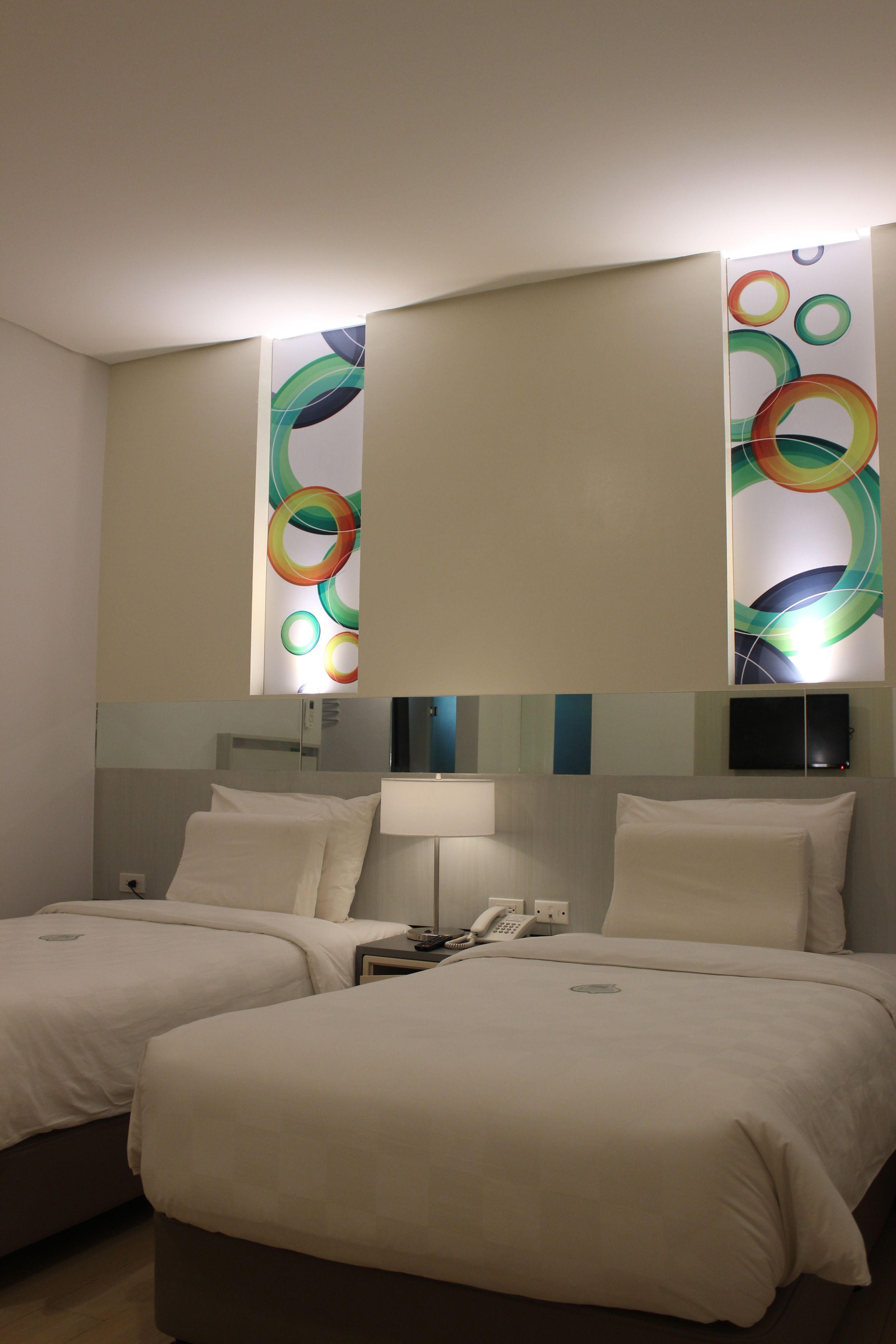 A hotel room with two beds, a bedside table with a lamp, and a wall-mounted TV.