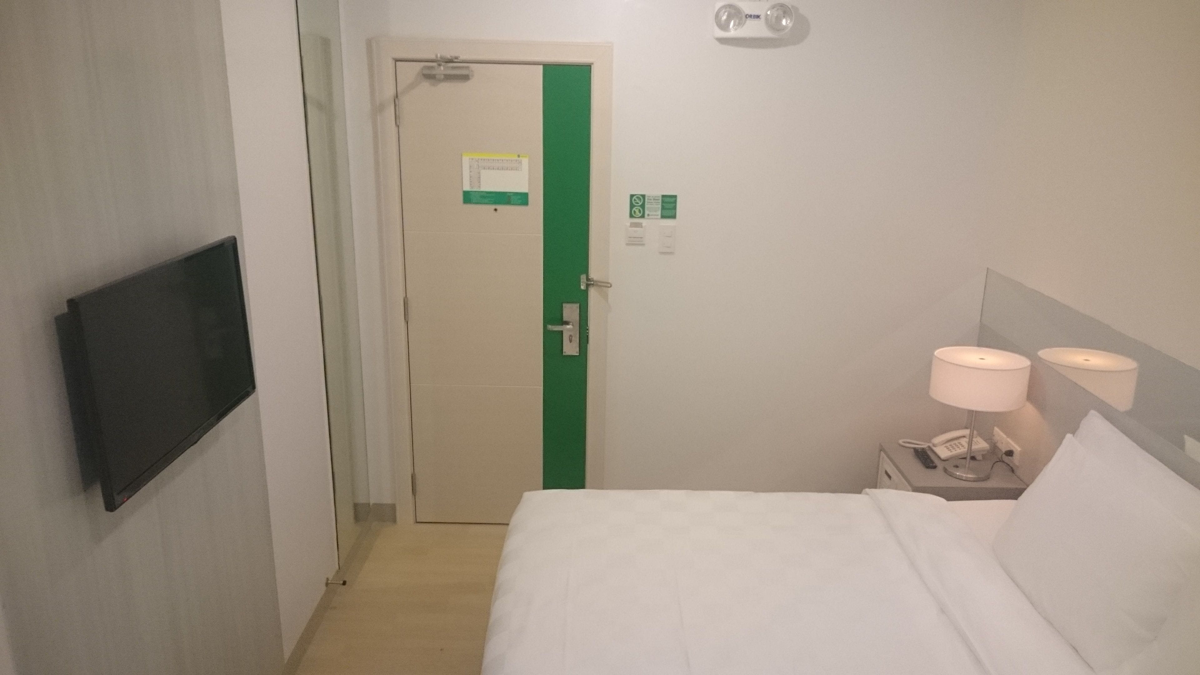 Quarto de hotel com uma cama, uma televisão fixada na parede, uma mesa de cabeceira com abajur e uma porta com faixa verde.