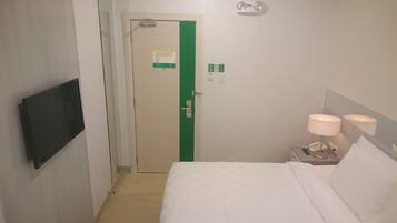 Quarto de hotel com uma cama, uma televisão fixada na parede, uma mesa de cabeceira com abajur e uma porta com faixa verde.