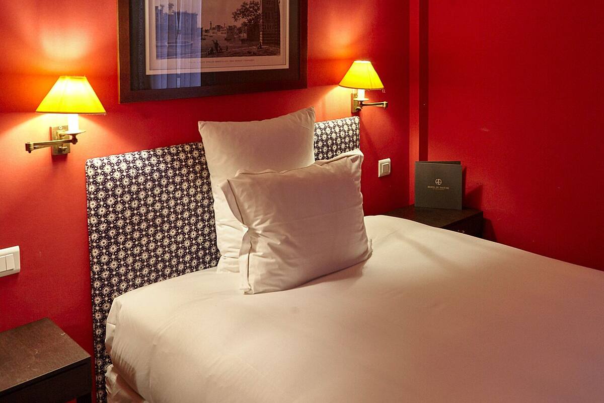 A hotel room with a red wall, a bed with white bedding, two bedside lamps, a framed picture on the wall, and a small table with a lamp.