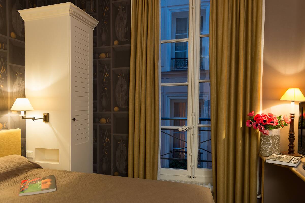 A hotel room with a bed, a nightstand with a lamp, a window with curtains, and a vase with flowers.