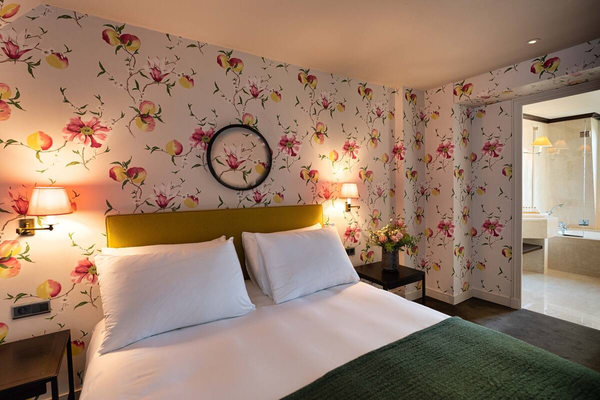 A hotel room with a bed, two bedside lamps, a round mirror, and a vase with flowers.