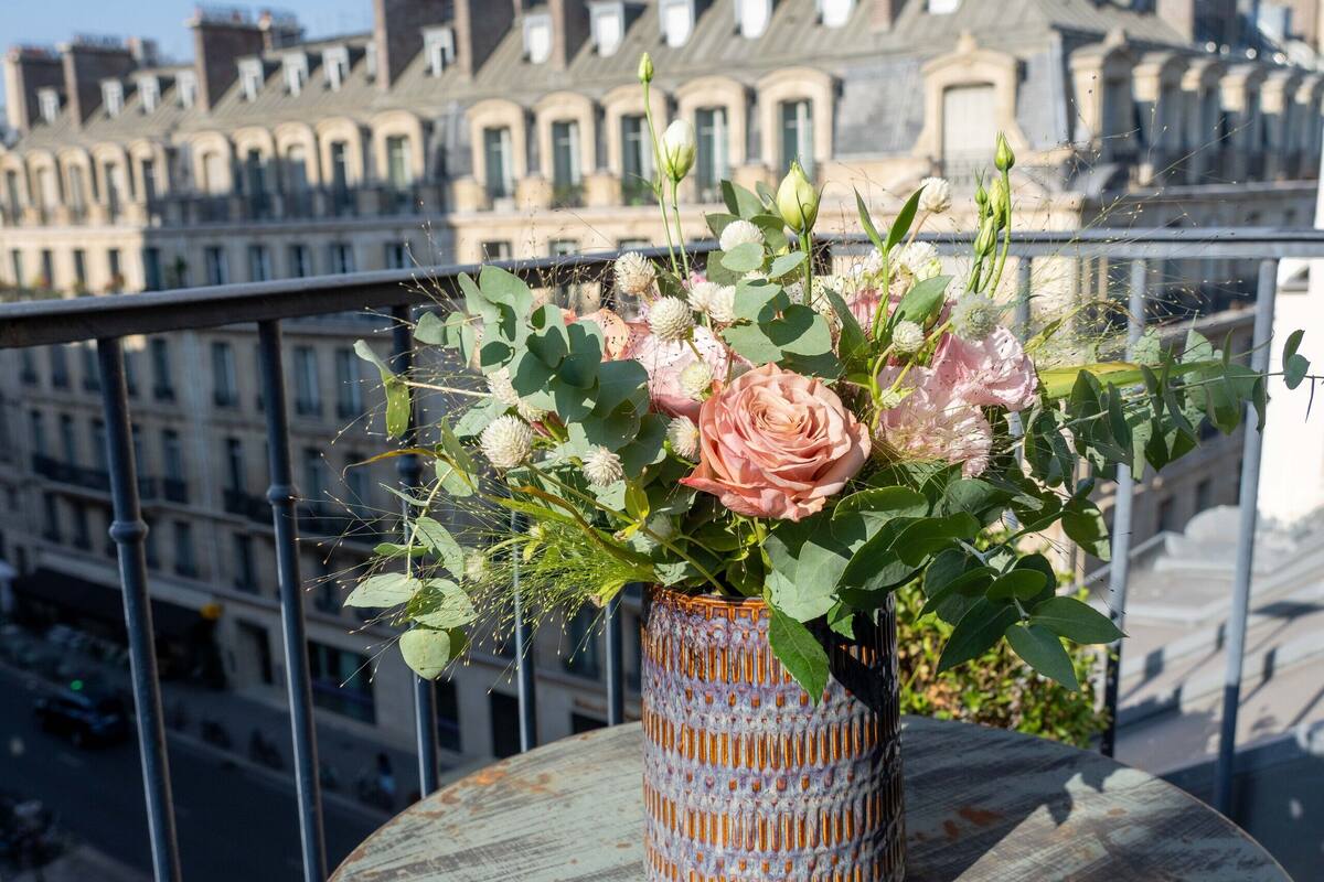 A balcony with a table holding a vase of flowers, overlooking a cityscape with buildings.