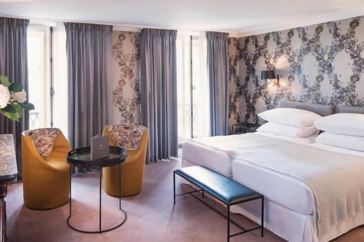 A hotel room with a bed, two armchairs, a small round table, a bench, a desk with a lamp, and a large window with curtains.