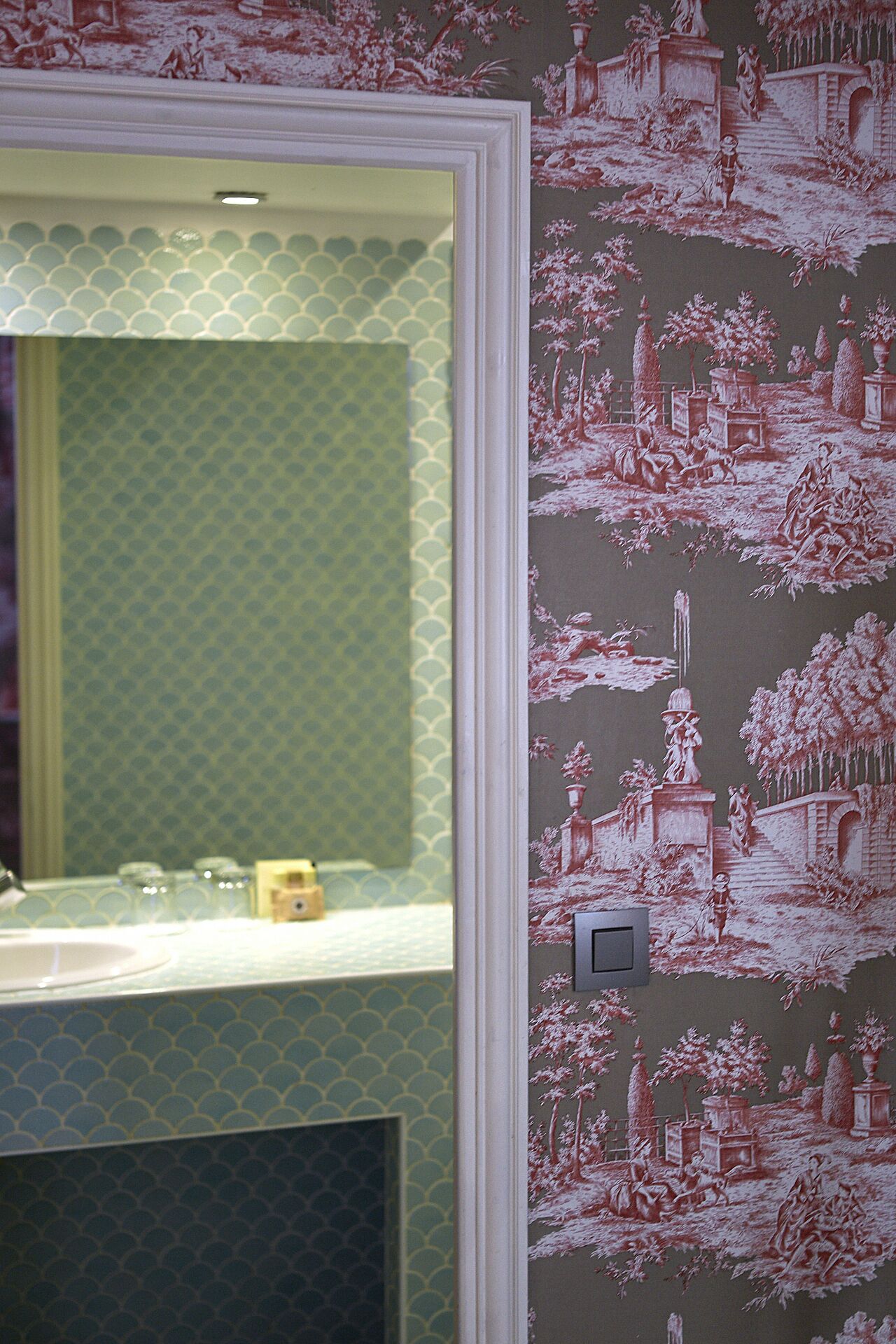 A bathroom with a mirror, a sink, and a decorative wallpaper featuring classical scenes.