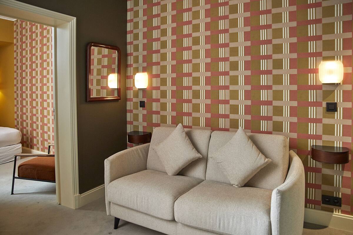 A neutral-colored sofa with two cushions in a room with patterned wallpaper and two wall-mounted lamps.