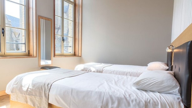 Un dormitorio con dos camas, suelo de madera y ventanas amplias con vistas a edificios.