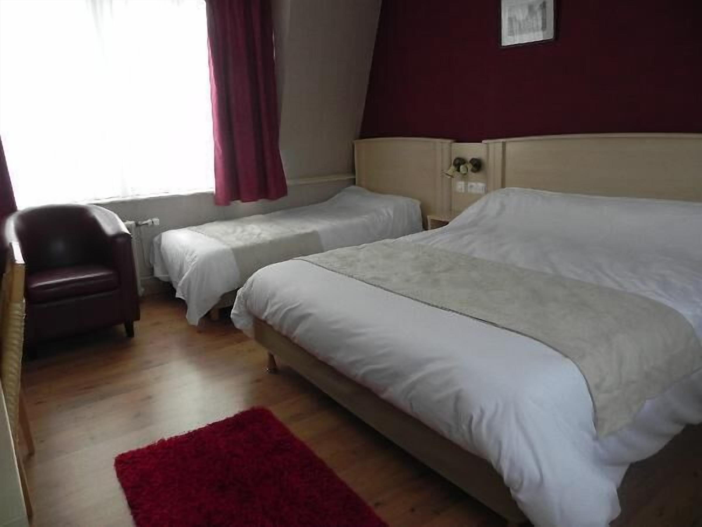 Un dormitorio con dos camas individuales, una alfombra roja, un sillón de cuero y una ventana con cortinas.