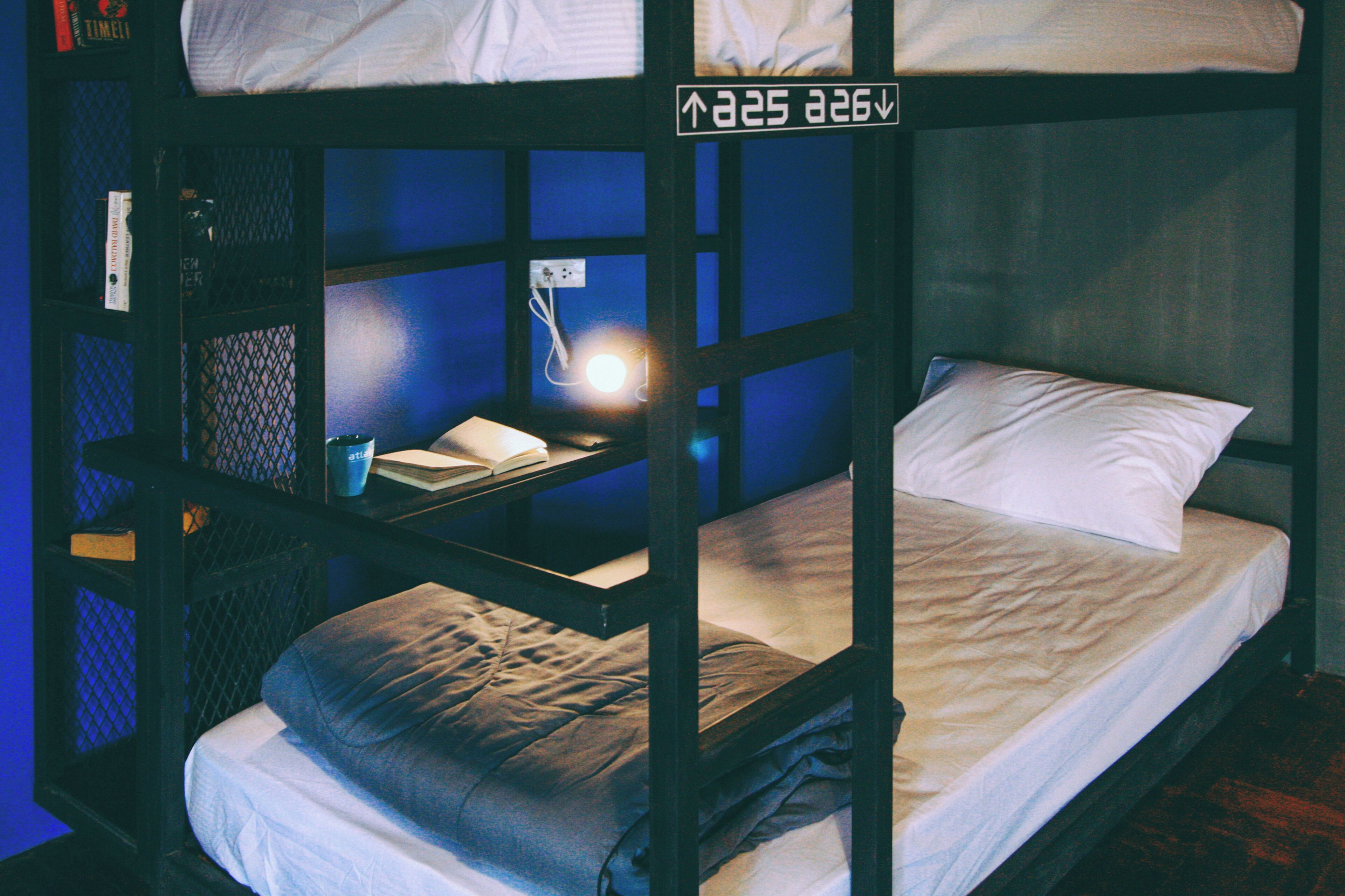 Una cama litera con una mesita pequeña y una lámpara, un almohadón blanco y un libro en la mesita de noche.