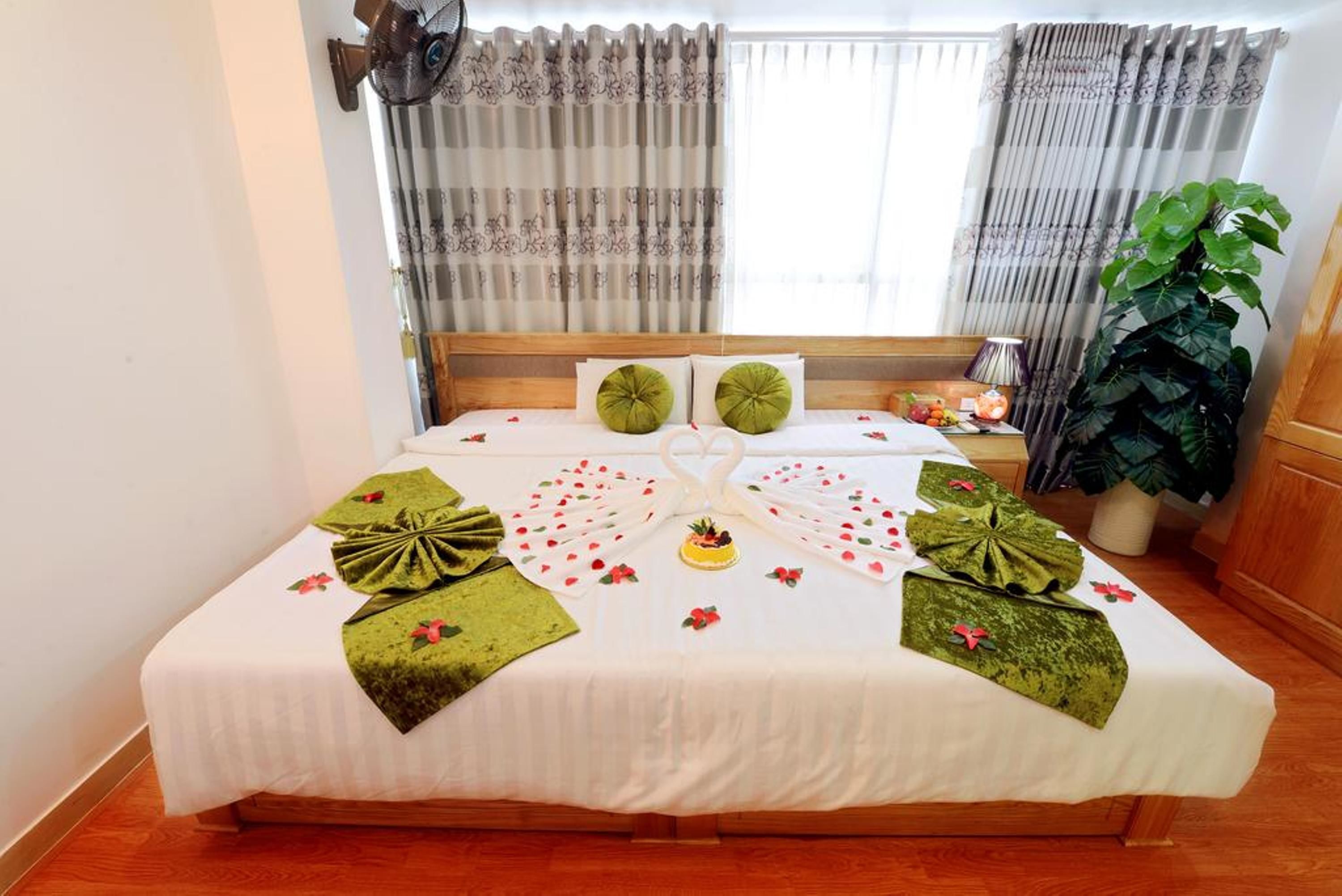 A neatly made bed with green towels and a fruit arrangement, a large potted plant, and a window with curtains.