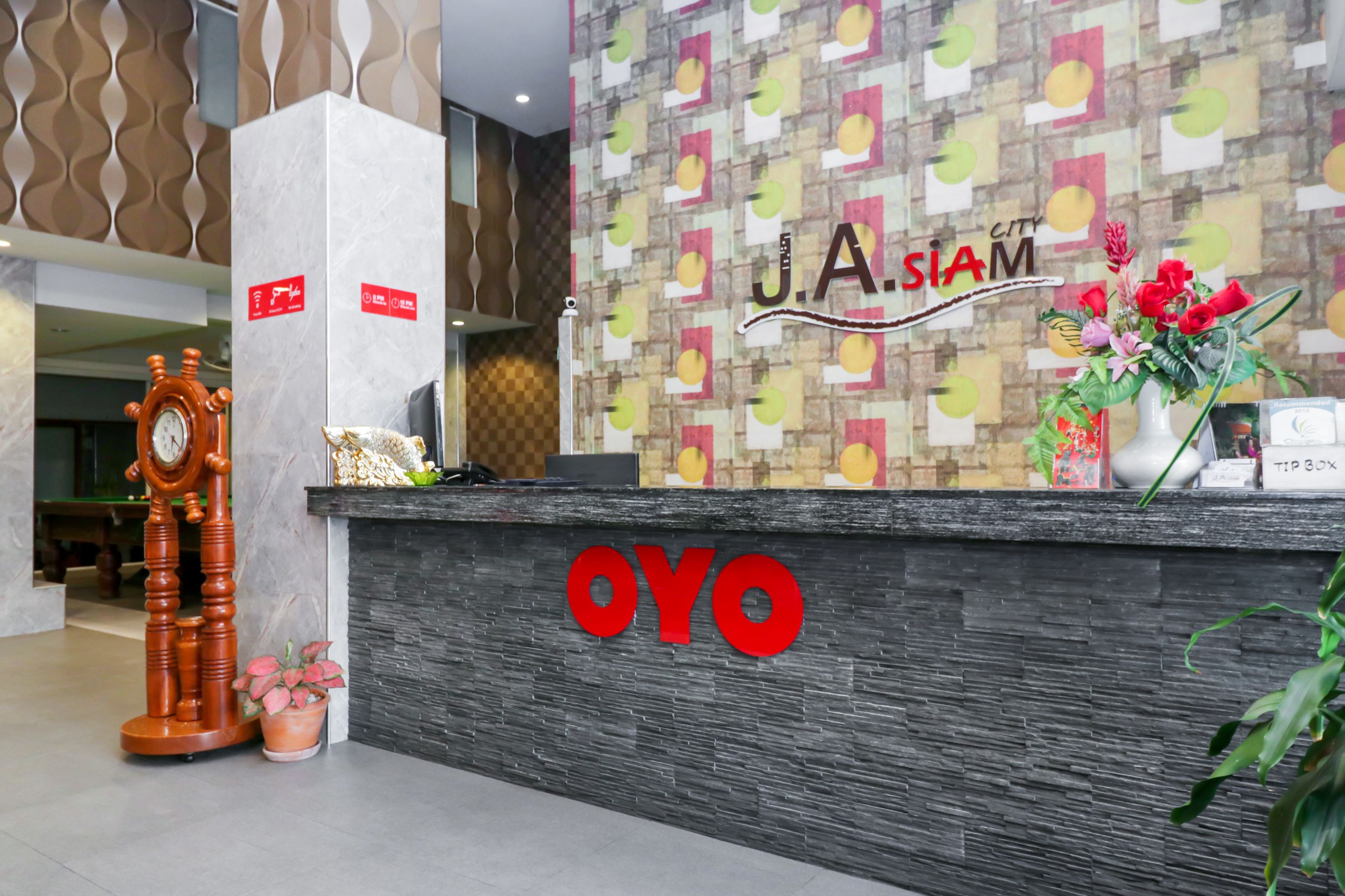A reception desk with a 'J.A.SIAM' sign and 'OYO' branding, a clock, and decorative items including a vase with flowers.