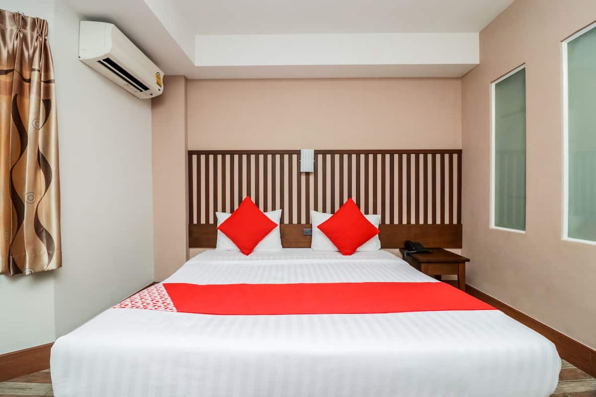 A hotel room with a large bed, a wooden headboard, a red accent pillow, a small bedside table, and a window.